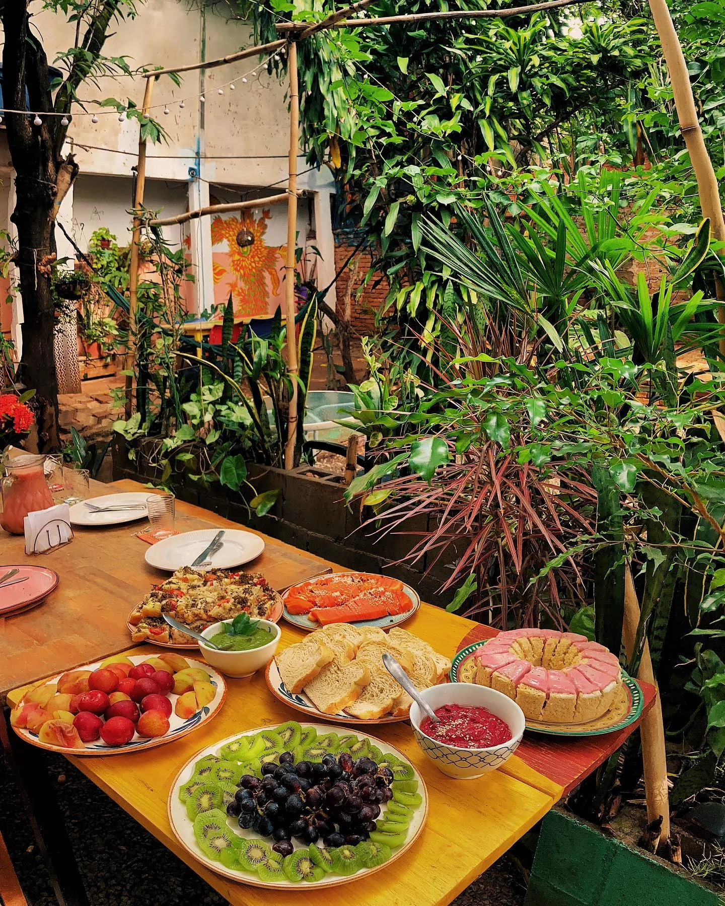 Mesa farta no pátio em dia claro — frutas, pães, doces e plantas ao redor