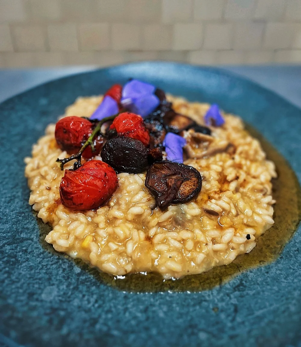 Risoto de milho verde em prato fundo, coberto com tomate cereja, shiitake e flores comestíveis