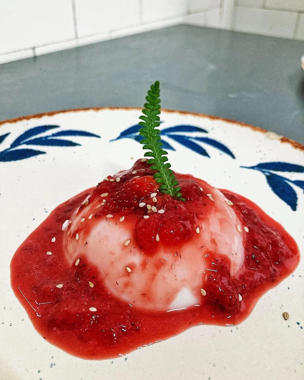 Panna cotta vegana com calda de frutas vermelhas e folhinha verde, em prato decorado azul e branco